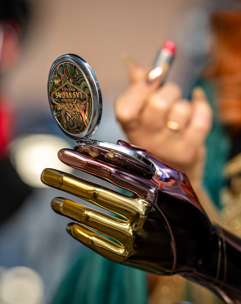 Zdjęcie metalicznej protezy ręki w odcieniach złota i fioletu. Aleksandra trzyma w niej lusterko, w drugiej ręce ma czerwoną pomadkę.