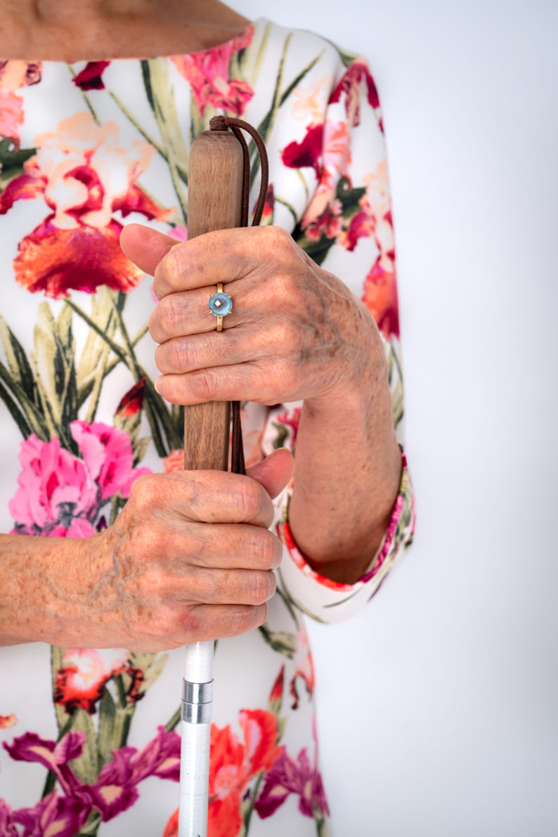 Zdjęcie dłoni Barbary trzymającej laskę dla osób niewidomych. Na lewej dłoni kobieta nosi złoty pierścionek z niebieskim kamieniem. Barbara ma sobie białą sukienkę z w kwiatowy wzór.