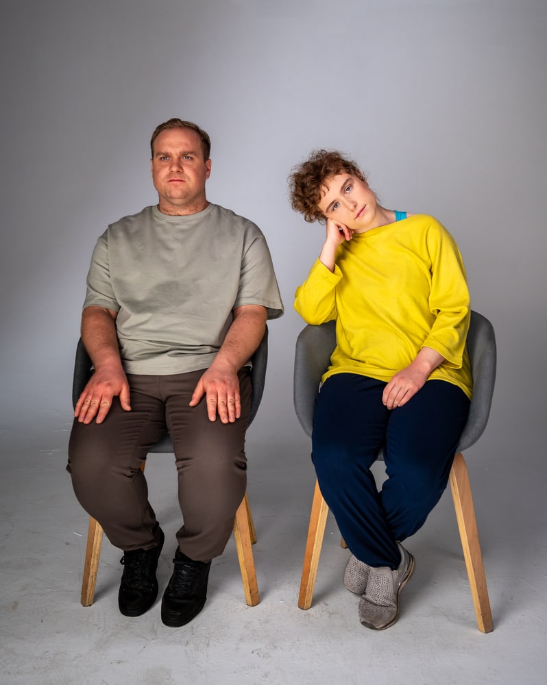 Zdjęcie Piotra i Marty z planu filmowego. Obydwoje siedzą na krzesłach na białym tle. W kadrze widać mikrofon. Marta podpiera głowę dłonią.