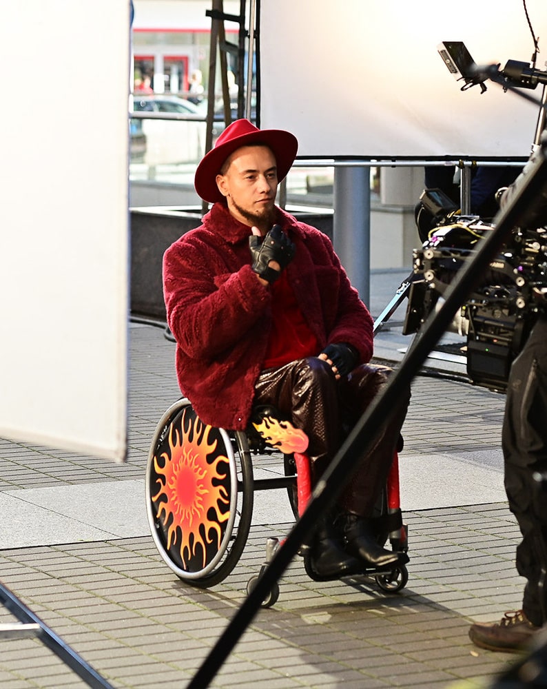 Zdjęcie Alana na planie zdjęciowym podczas ujęcia. Mężczyzna porusza się na wózku, jest ubrany w bordowe ekstrawaganckie futro oraz czerwoną koszulę, na głowie ma bordowy kapelusz z dużym rondem. Patrzy w bok w kamerę filmową, poprawia na dłoniach czarne, skórzane rękawiczki bez palców.