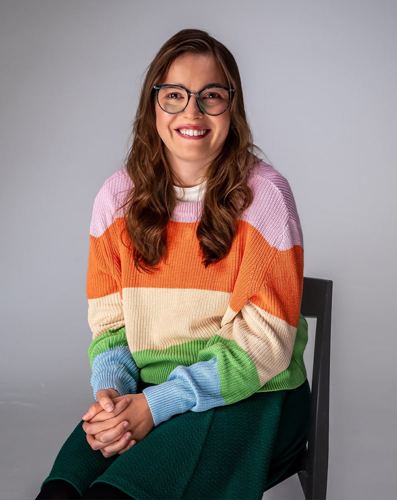 Portret Julii patrzącej w obiektyw z uśmiechem. Dziewczyna ma na sobie sweter w pasy w kolorach: różowym, pomarańczowym, żółtym i zielonym. Nosi okulary korekcyjne