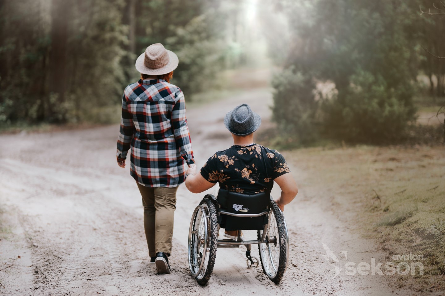 Na zdjęciu kobieta i mężczyzna na wózku podczas spaceru. Są odwróceni tyłem i trzymają się się za ręce.