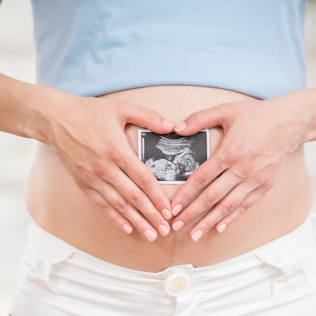 Na zdjęciu brzuch kobiety w ciąży, na nim ręce w których jest zdjęcie z USG.