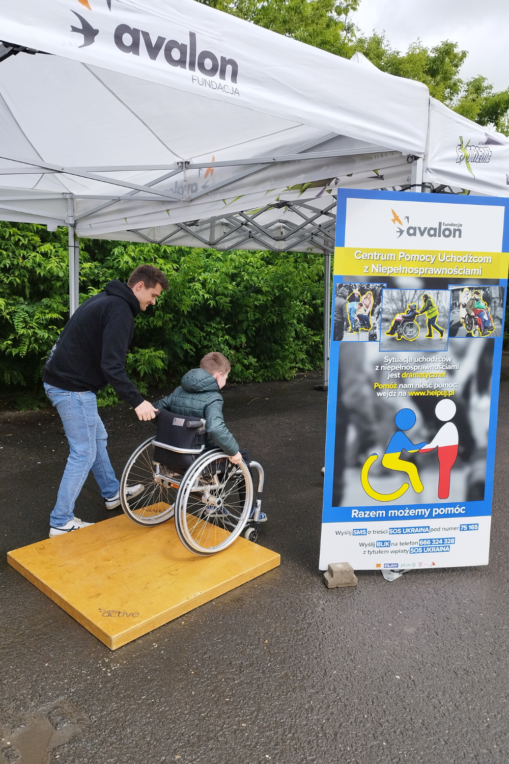 Na zdjęciu fizjoterapeuta pomaga chłopcu pokonać tor przeszkód na wózku. Obok rool-up Centrum Pomocy Uchodźcom z Niepełnosprawnościami