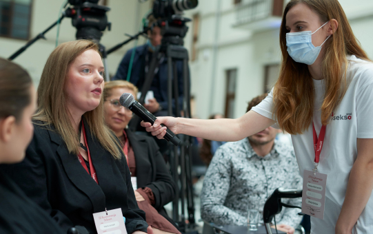 Jakiś podpis Na zdjęciu wolontariuszka podaje mikrofon osobie wypowiadającej się.