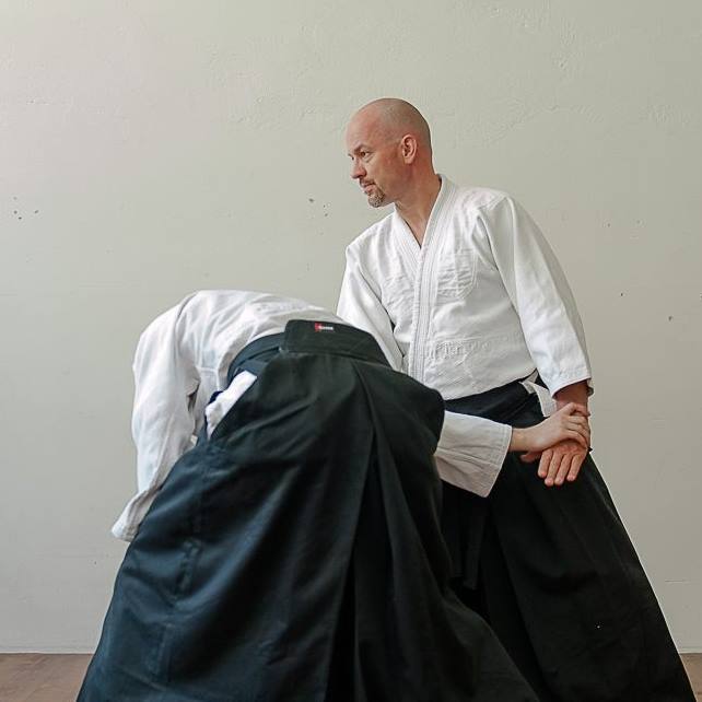 Na zdjęciu dwóch mężczyzn ćwiczacych aikido - jeden stoi wyprostowany, drugi się pochyla i trzyma pierwszego za rękę.