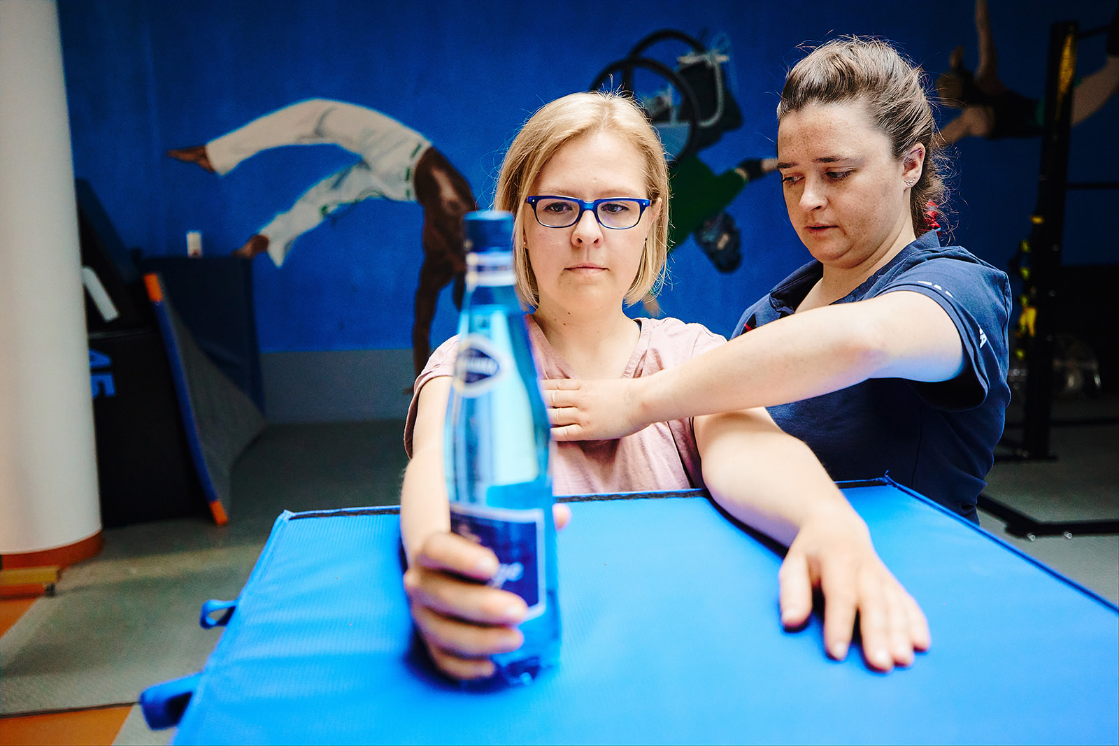 Na zdjęciu fizjoterapeutka i pacjentka podczas zajęć przy leżance. Pacjenta ma wyciągnięte ręce przed siebie, w jednej trzyma butelkę wody.