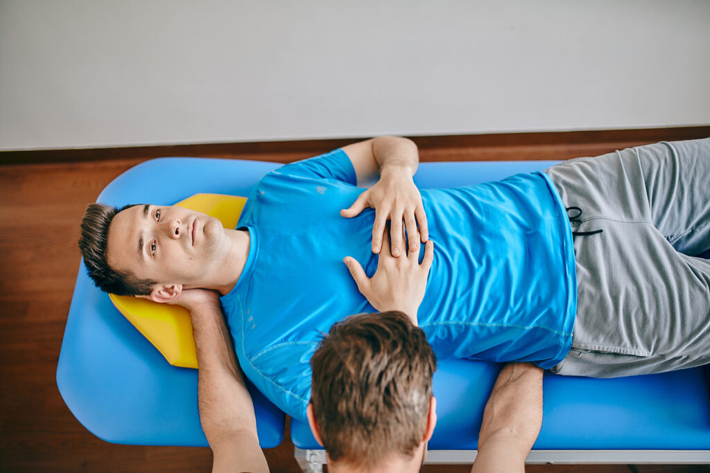 Na zdjęciu fizjoterapeuta trzymający ręce pod plecami pacjenta lezącego na leżance.