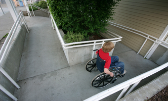 Na zdjęciu chłopiec na wózku zjeżdża z podjazdu przy budynku.