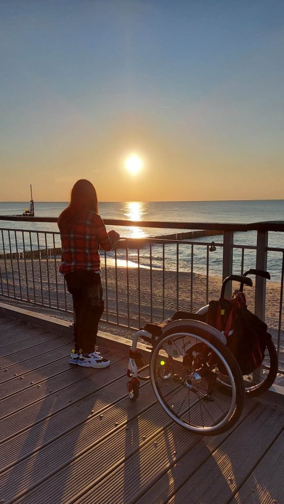 Młoda dziewczyna obserwuje zachód słońca na plaży. Obok niej, na pomoście, stoi wózek. 