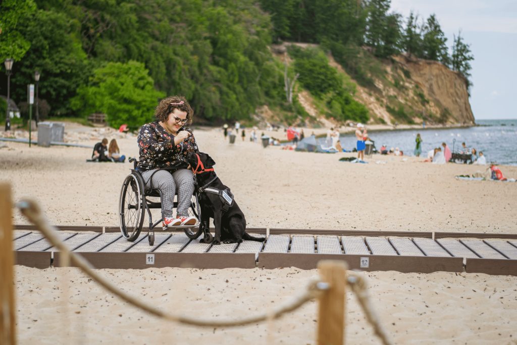 Uśmiechnięta dziewczyna na wózku głaska psa. Znajdują się na plaży.