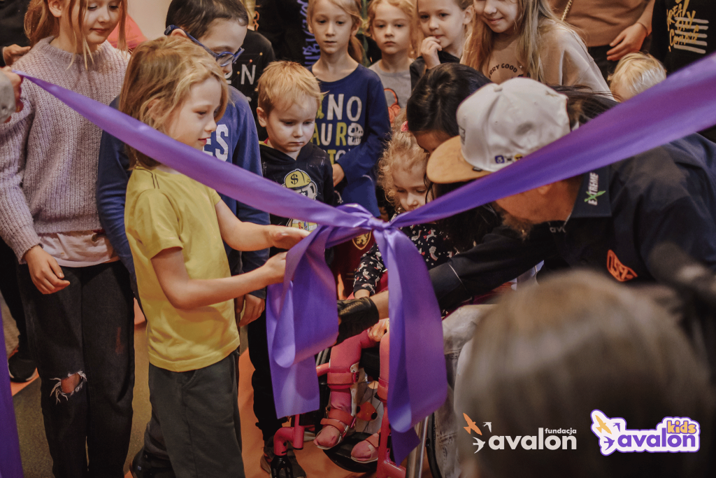 Prezes Fundacji Avalon przecina wstęgę przy pomocy małej dziewczynki. Otacza ich duża grupa dzieci.