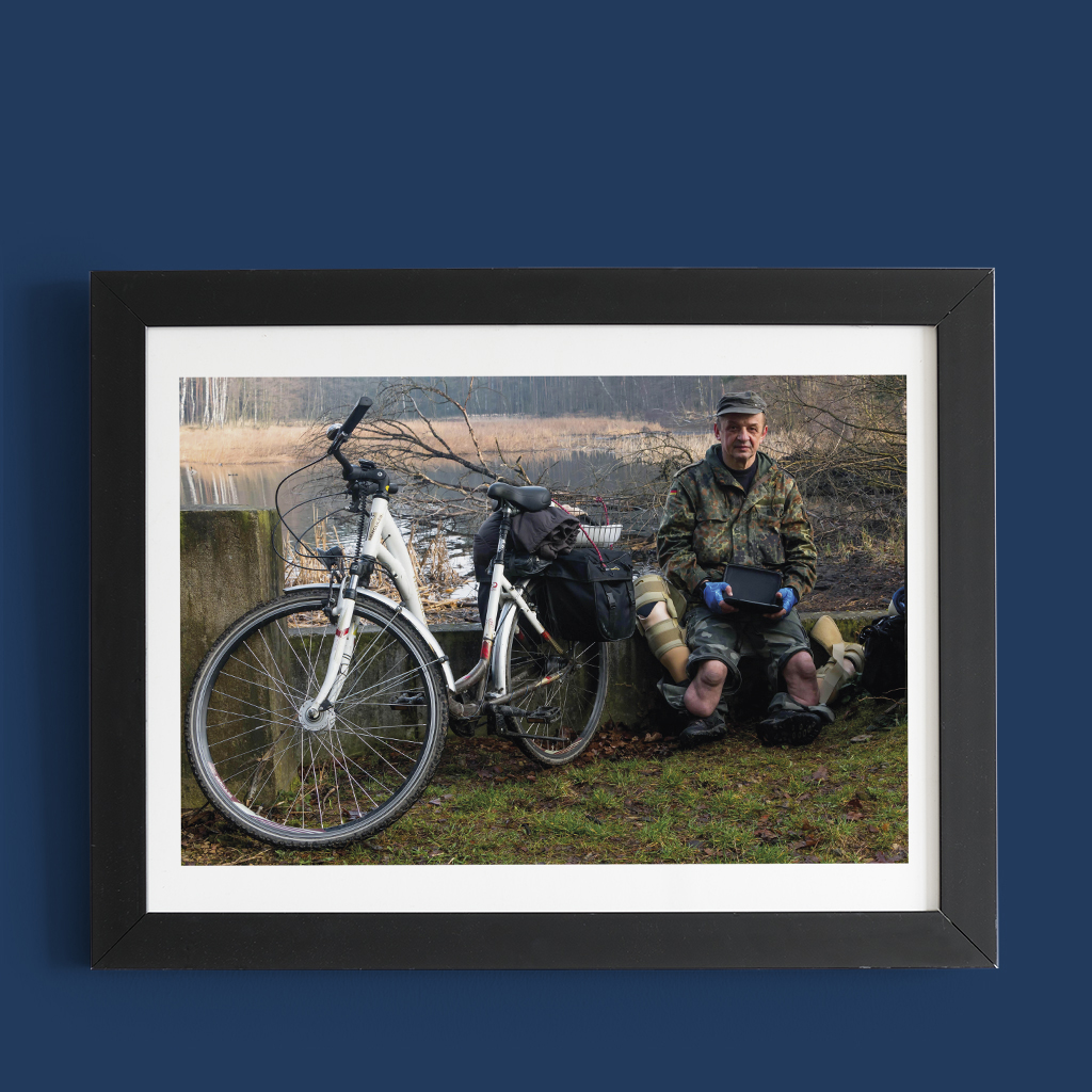 Mężczyzna bez nóg w mundurze wojskowym siedzi nad jeziorem, obok znajduje się rower.