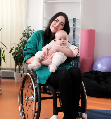 Zdjęcie kobiety na wózku trzymającej na kolanach kilkumiesięczną córeczkę. Są one w sali rehabilitacyjnej, w tle przyrządy do ćwiczeń.