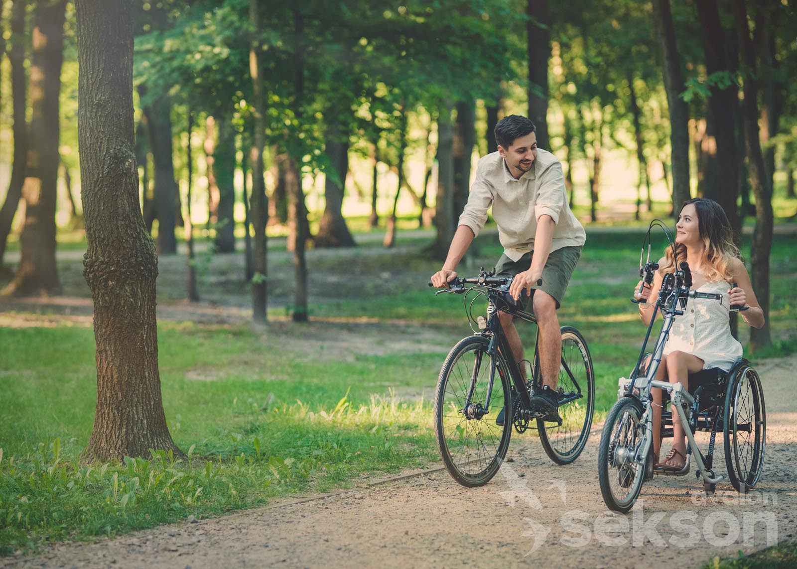 Karol z Julią na rowerach jadą przez park. Julia jedzie na rowerze dostosowanym do jej wózka inwalidzkiego.