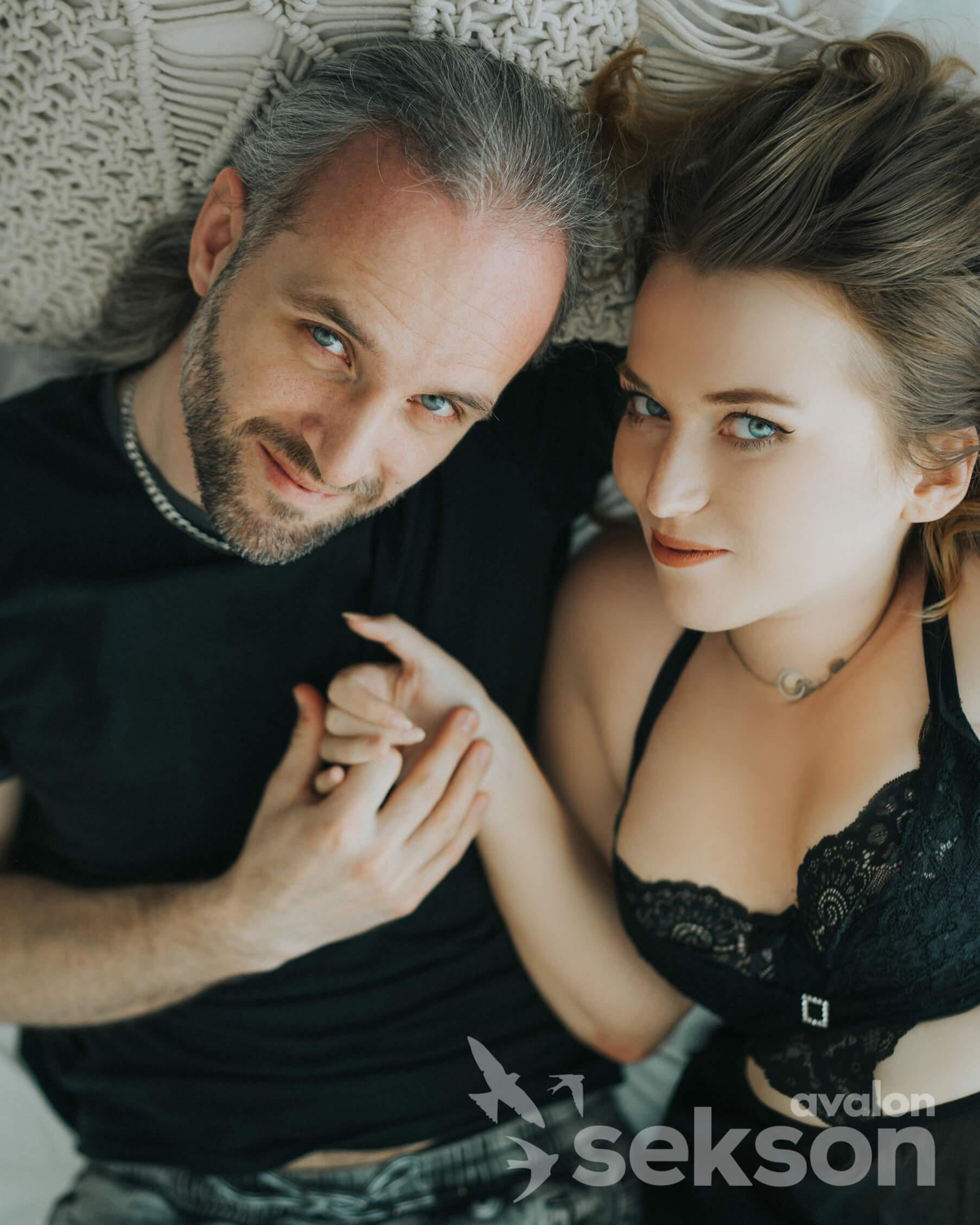 Sylwia i Marcin widoczni od popiersia, leżą na łóżku. Trzymają się za ręce i patrzą w obiektyw.