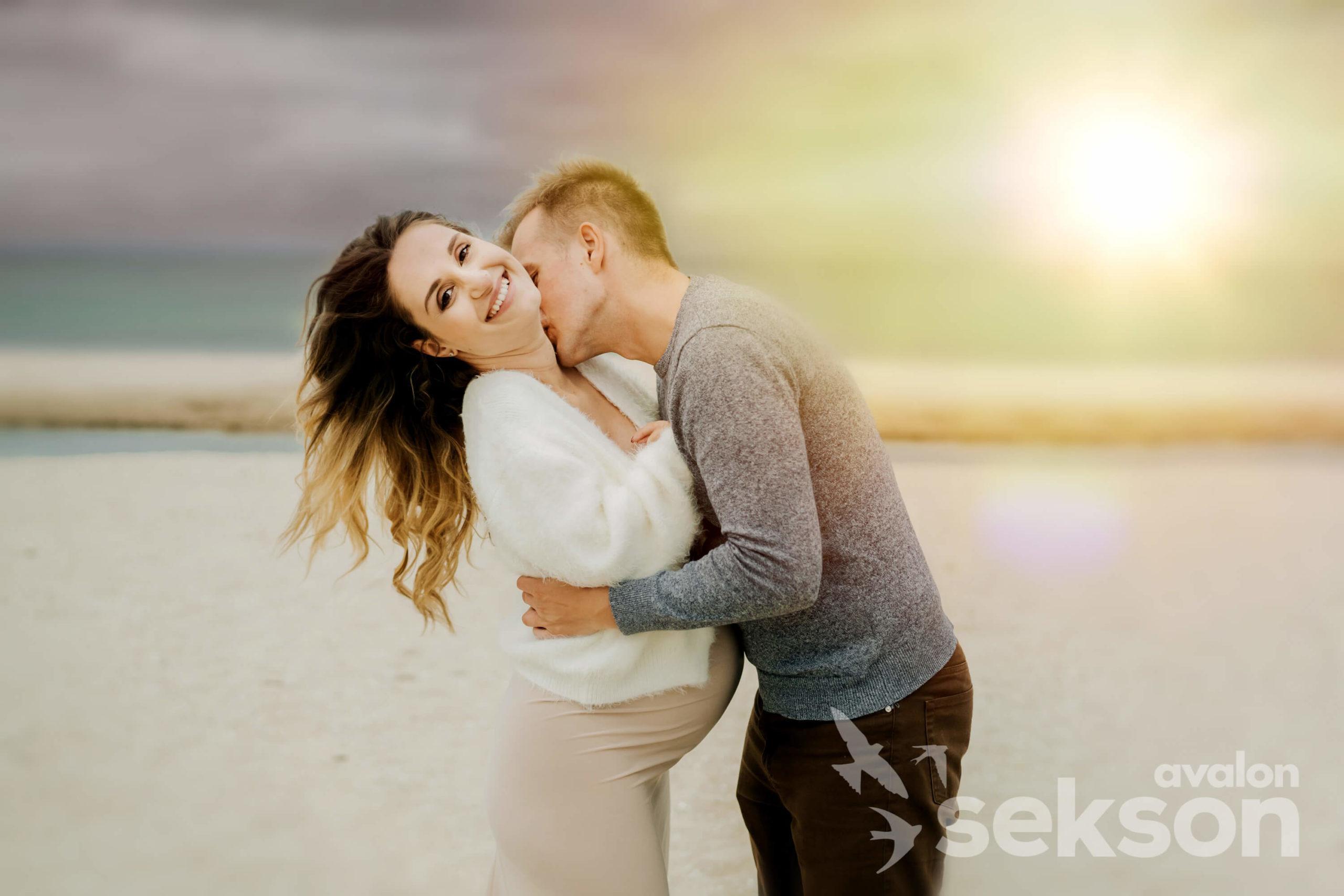 Sylwia i Tomasz na plaży. Tomasz całuje Sylwię w szyję. W tle zachód słońca.