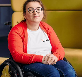 Na zdjęciu uśmiechnięta dziewczyna na wózku. Widzimy ją od kolan w górę. Ma na sobie okulary z czarnymi oprawkami, niebieskie dżinsy, pomarańczowy sweterek i białą koszulkę z niedużym logotypem fundacji avalon.