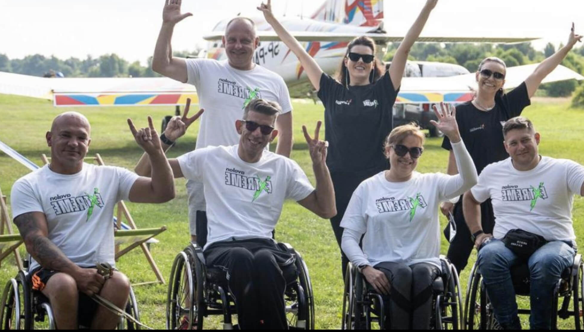 grupa 4 osób na wózkach pozuje do zdjęcia - wszyscy ubrani są w białe koszulki z logotypem Avalon Extreme; za nimi stoi trener i kierowniczki projektu Avalon Extreme; w tle tereny zielone