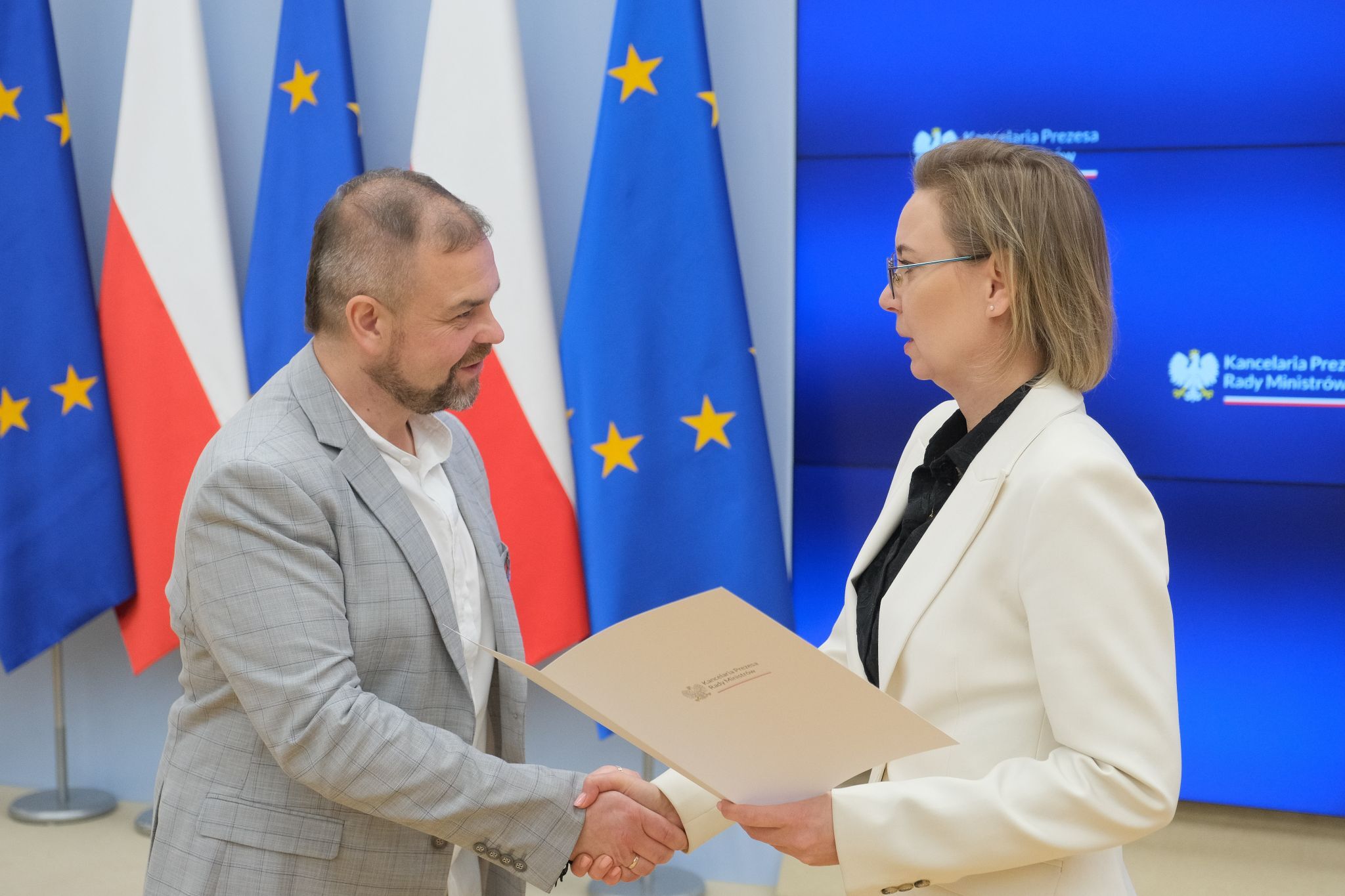 Łukasz Wielgosz ściska rękę kobiety przekazującej mu teczkę.