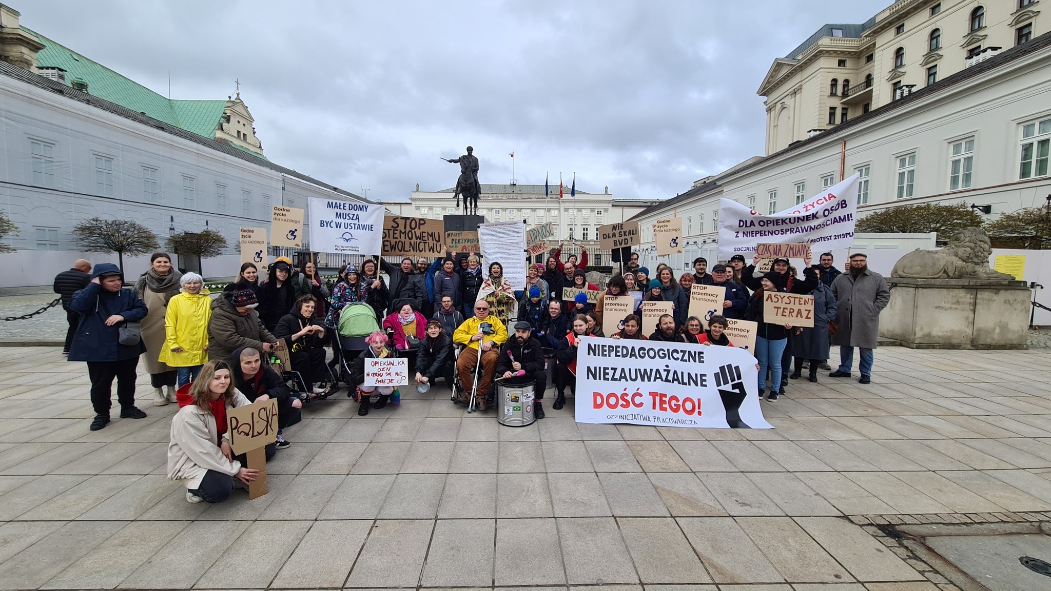 Grupa osób pełnosprawnych i z niepełnosprawnościami z transparentami przed Pałacem Prezydenckim w Warszawie.