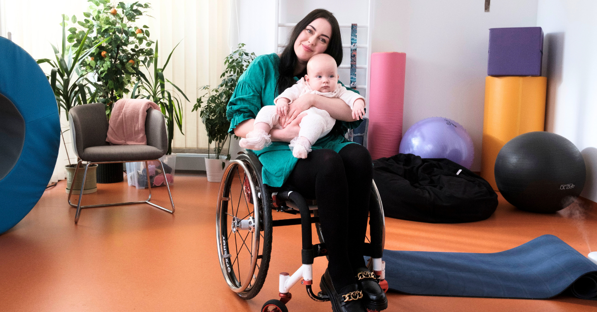 Kobieta na wózku z niemowlakiem na rękach. Znajdują się w sali rehabilitacyjnej.