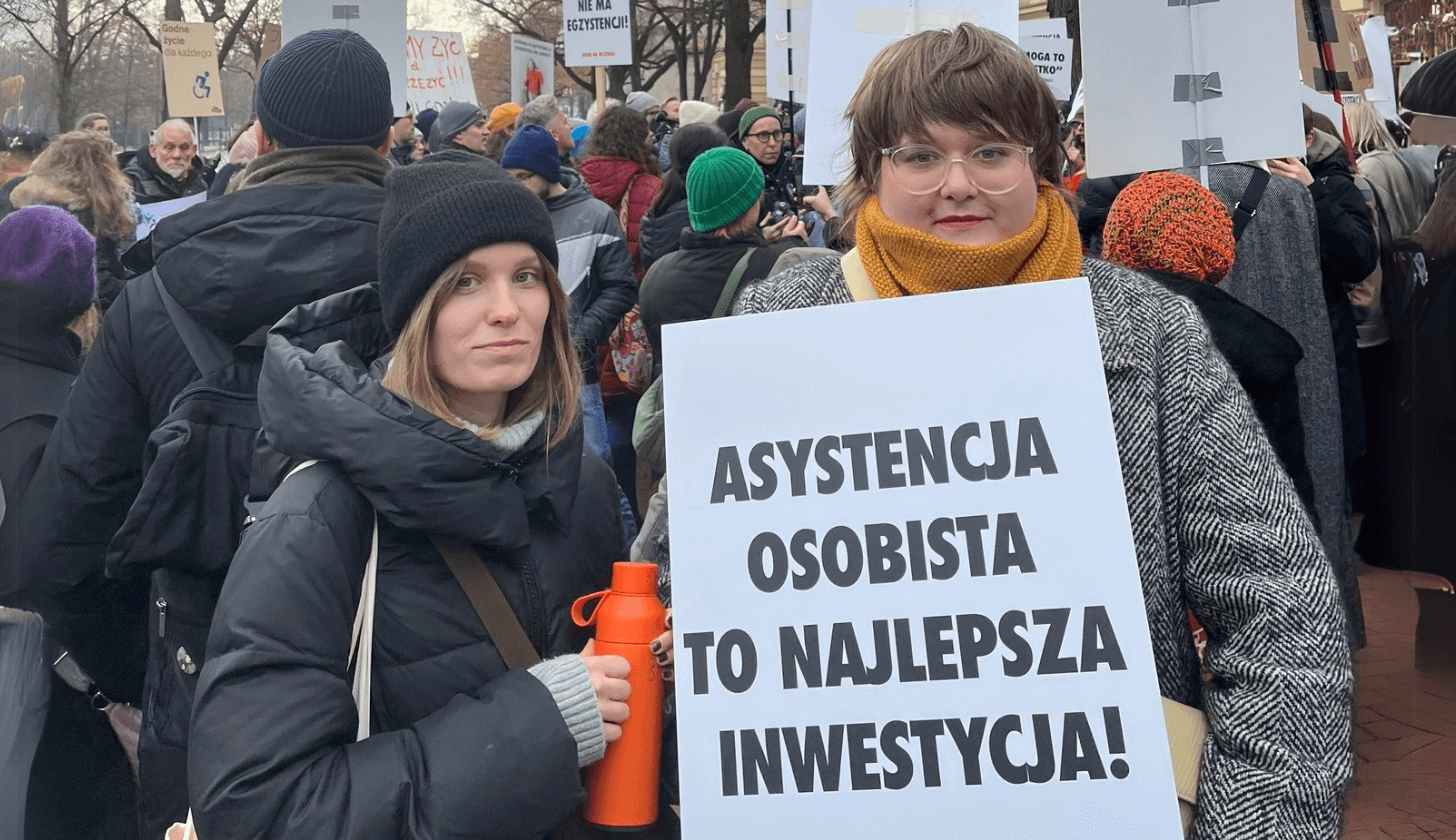 Dwie młode kobiety podczas strajku. Są ubrane w kurtki i trzymają transparent z napisem Asystencja osobista to najlepsza inwestycja.