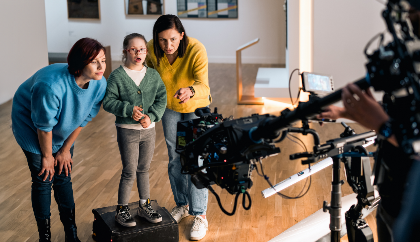 Ekipa filmowa nagrywa scenę w galerii sztuki – w kadrze widać dwie kobiety i dziecko oglądające obrazy, a w tle sprzęt filmowy oraz operator kamery
