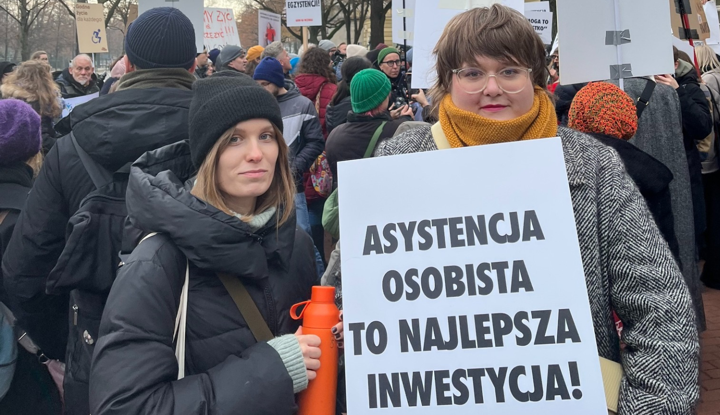 Dwie osoby w trakcie protestu, trzymają baner z napisem: