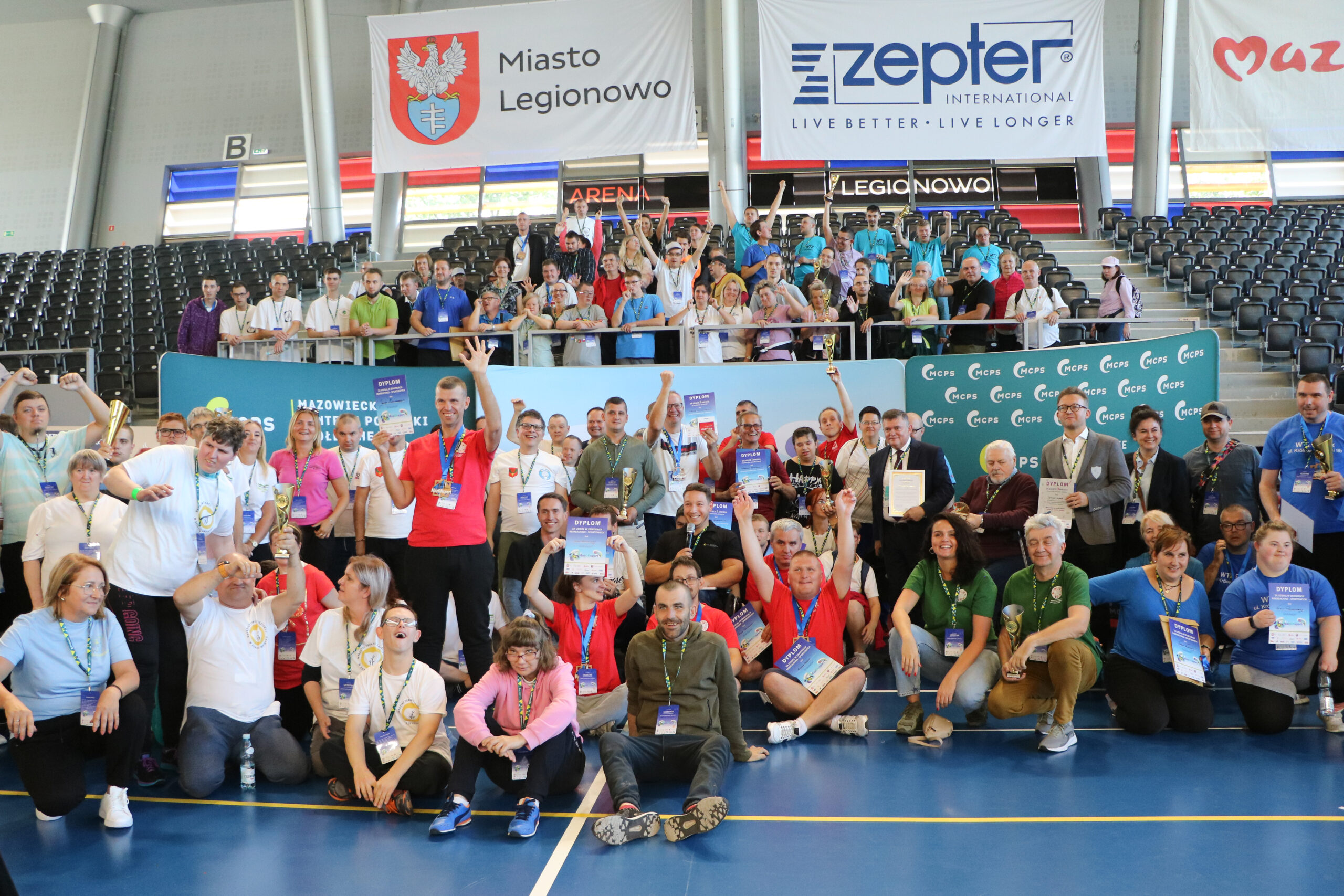 Zdjęcie z pierwszych zawodów MazONsport, przedstawia uczestników i organizatorów, którzy pozują do wspólnego zdjęcia grupowego na Arenie Legionowo, w tle trybuny.