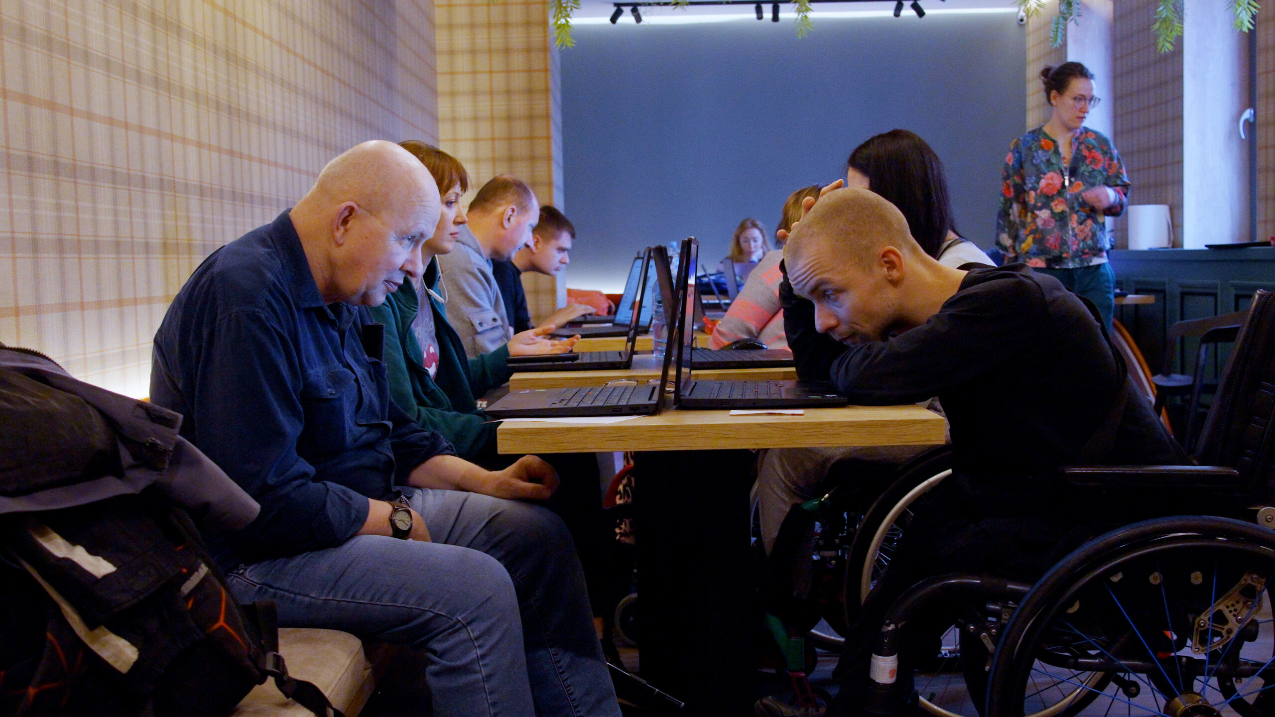 Grupa osób siedzi przy długim stole w pomieszczeniu o ciepłym, przytulnym wystroju. Każdy pracuje przy własnym laptopie. Po lewej stronie starszy mężczyzna w niebieskiej koszuli skupiony jest na ekranie. Po prawej stronie młody mężczyzna na wózku inwalidzkim opiera głowę na rękach, patrząc na monitor. W tle widać kolejne osoby zaangażowane w pracę oraz kobietę stojącą w kwiecistej bluzce, obserwującą grupę. Atmosfera jest skupiona i spokojna.