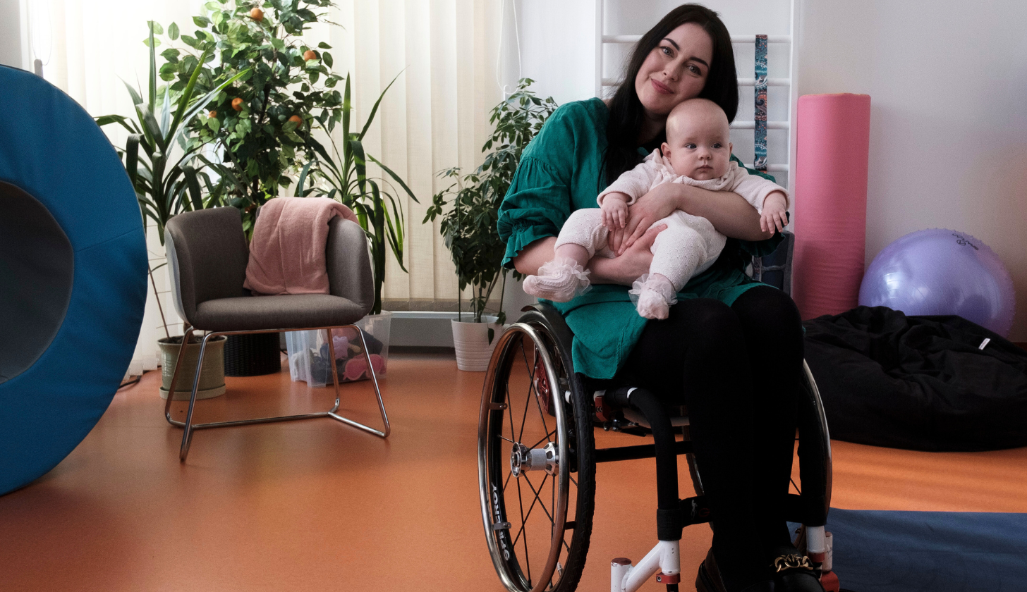 Młoda kobieta na wózku inwalidzkim siedzi w jasnym, przytulnym pomieszczeniu i trzyma na kolanach niemowlę. Oboje patrzą z uśmiechem w stronę aparatu. W tle znajdują się rośliny doniczkowe, fotel z różowym kocem, sprzęt rehabilitacyjny i piłki do ćwiczeń.