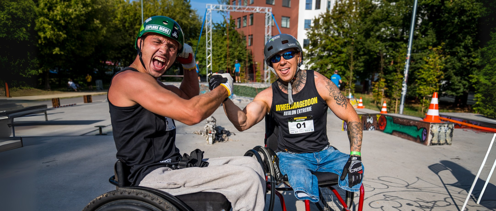 Dwóch uczestników zawodów Wheelmageddon na wózkach ściska sobie dłonie. Obaj mają na sobie sportowe koszulki na ramiączkach, ochraniacze i kaski. Jeden z nich krzyczy z entuzjazmem, drugi uśmiecha się szeroko.