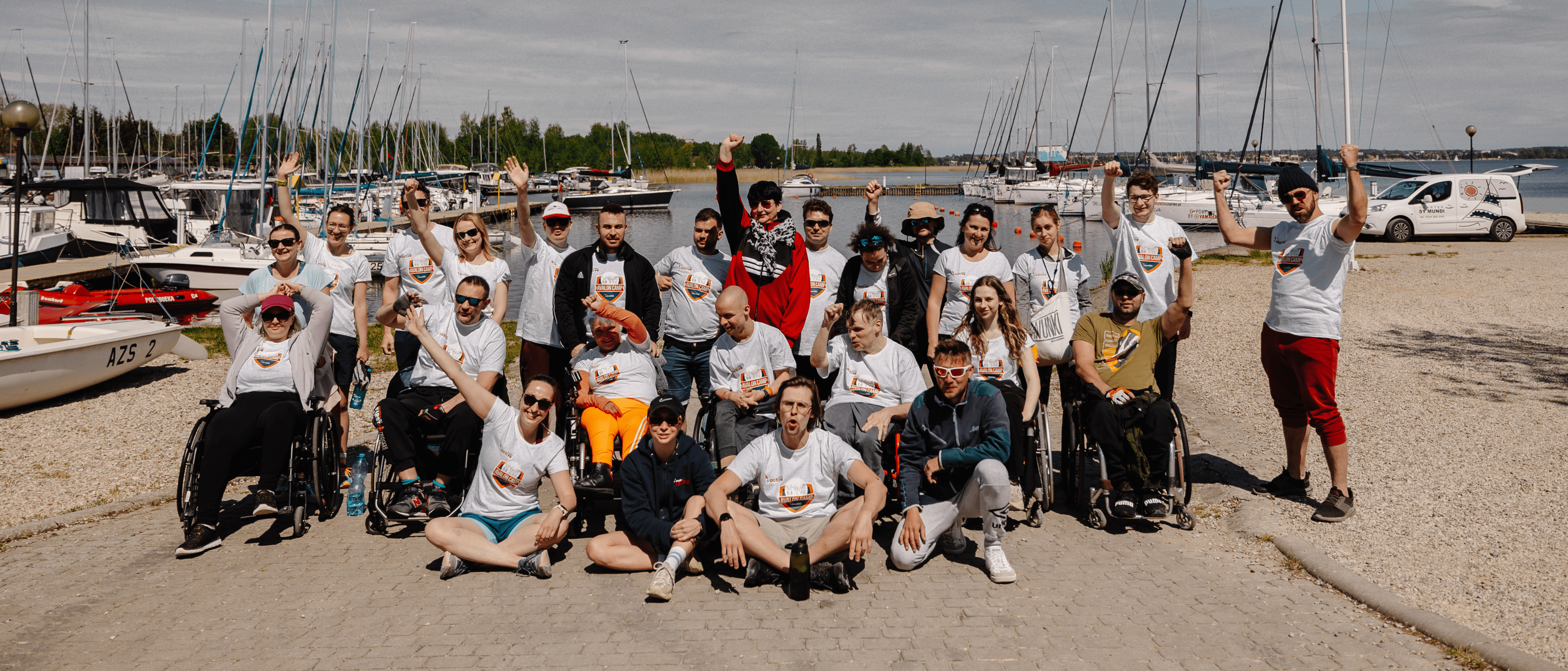 Uczestnicy Avalon Camp nad brzegiem jeziora. Za nimi łodki w porcie.