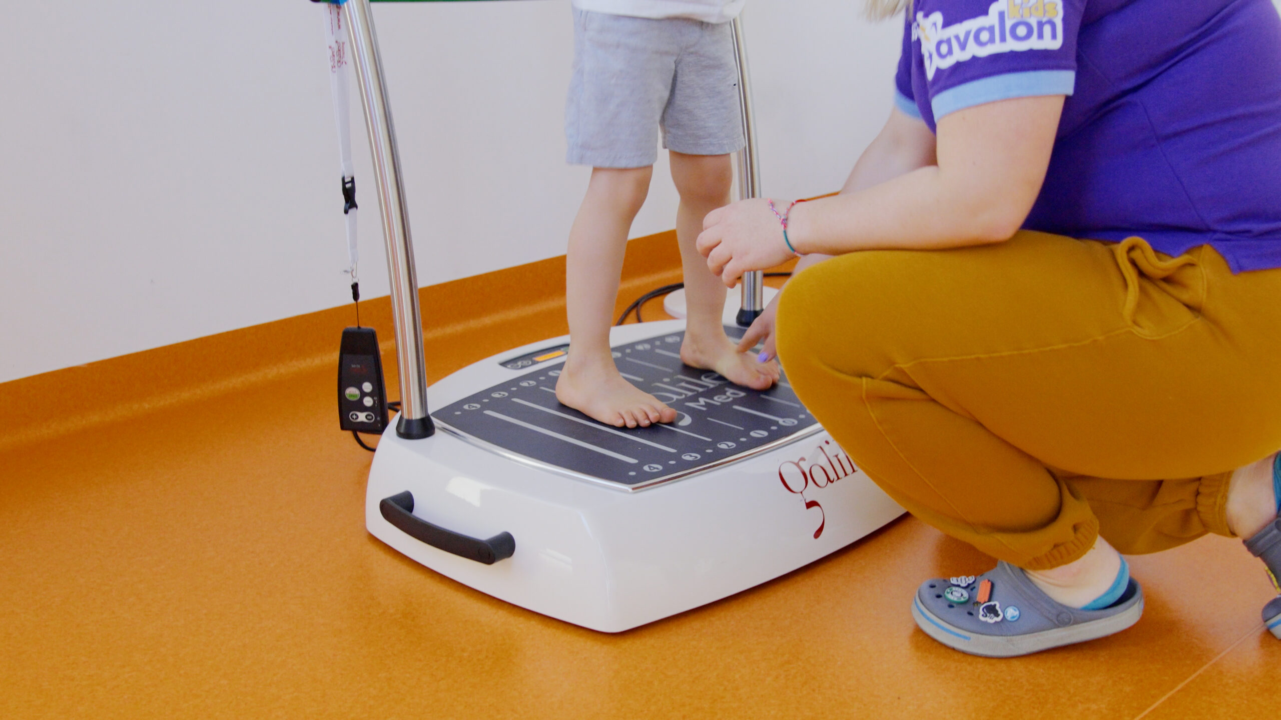 Chłopiec stoi bosymi stopami na urządzeniu do stymulacji. Obok jest fizjoterapełta.