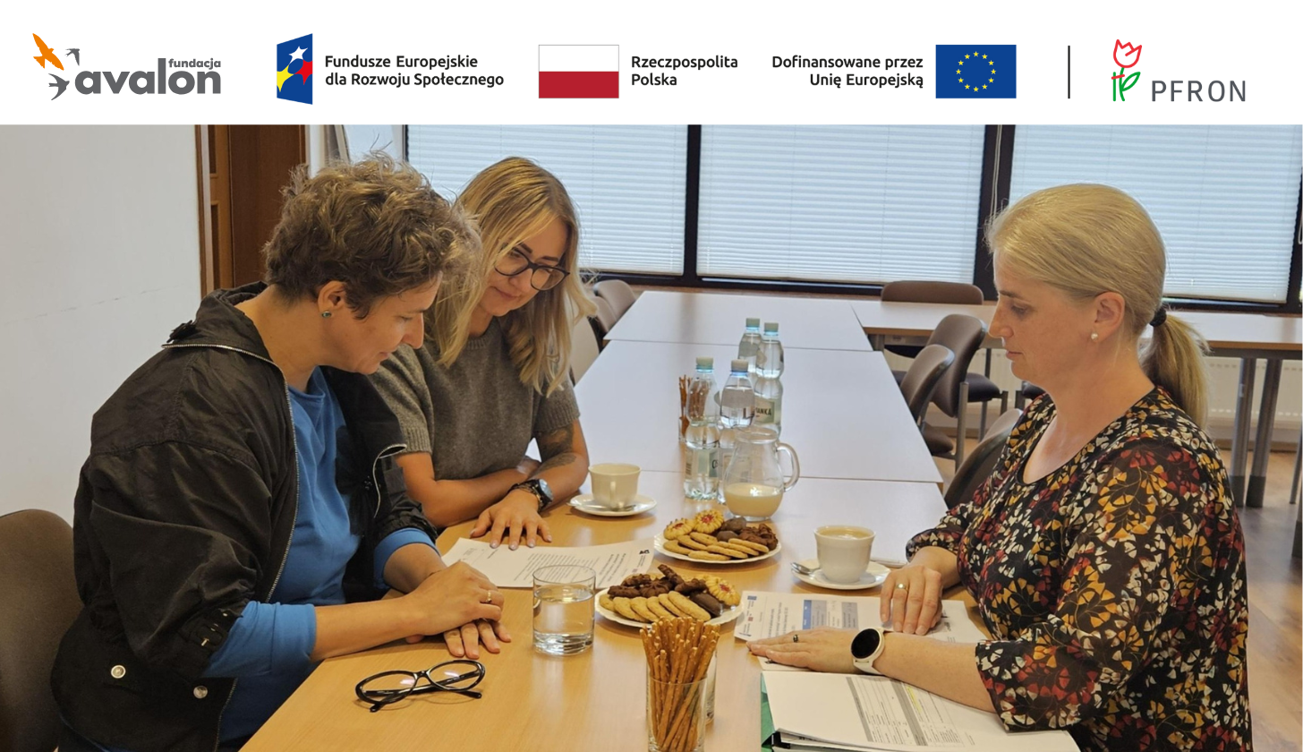 Na zdjęciu widać trzy kobiety siedzące przy długim stole w sali konferencyjnej. Na stole znajdują się dokumenty, szklanki z wodą, filiżanki z kawą lub herbatą oraz talerzyki z ciastkami. Kobiety skupiają się na dokumentach leżących przed nimi. Nad nimi widoczne są logotypy Fundacji Avalon, Funduszy Europejskich, Rzeczypospolitej Polskiej oraz Unii Europejskiej, a także logo PFRON.