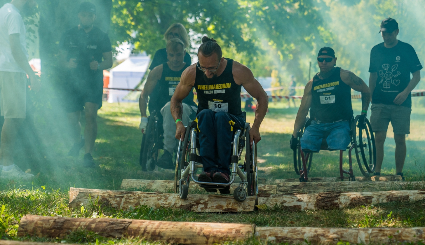 Osoba na wózku ubrana w czarną koszulkę z żółtym napisem Wheelmageddon pokonuje tor przeszkód podczas wydarzenia. W tle inni zawodnicy.