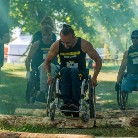 Osoba na wózku ubrana w czarną koszulkę z żółtym napisem Wheelmageddon pokonuje tor przeszkód podczas wydarzenia. W tle inni zawodnicy.