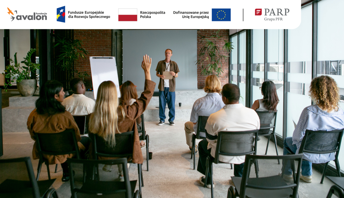 Grupa osób w sali warsztatowej siedzi na krzesłach. Jedna z kobiet podnosi rękę. Prowadzący szkolenie stoi przed grupą, obok flipcharta.