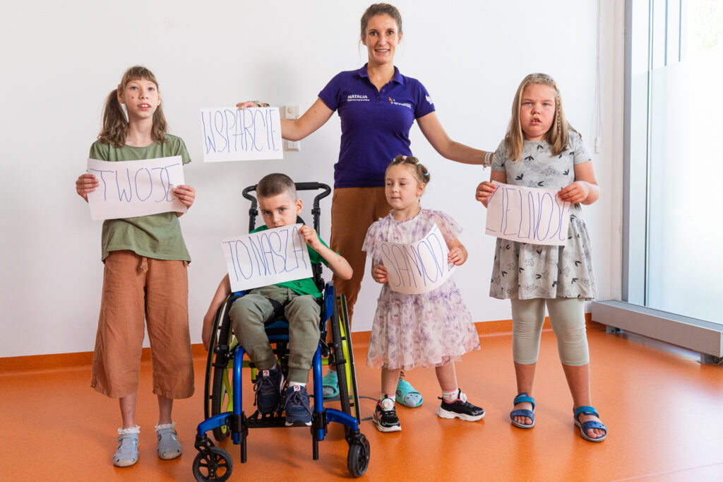 Czwórka dzieci z niepełnosprawnościami i fizjoterapeutka. Trzymają kartki na których jest napisane: Twoje wsparcie to nasza samodzielność.
