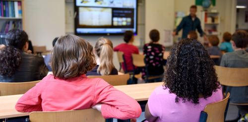 Dzieci siedzą w klasie i sluchają nauczyciela. Widzimy ich tyłem.