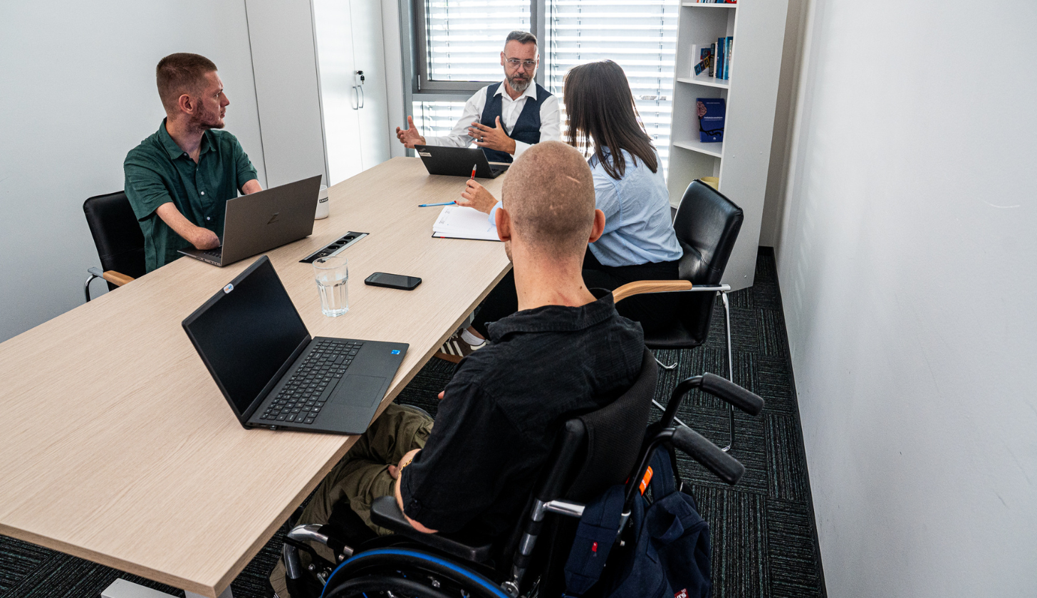 Zdjęcie przedstawia cztery osoby uczestniczące w spotkaniu przy długim stole w jasnej sali konferencyjnej. Dwóch mężczyzn i jedna kobieta siedzą przy stole, rozmawiając i pracując na laptopach. Czwarta osoba, mężczyzna, siedzi tyłem do obiektywu na wózku.