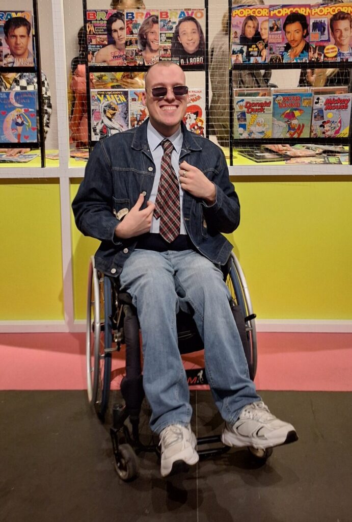 Norbert Tumidaj na wózku w dżinsowych spodniach, dżinsowej marynarce,, białej koszuli i krawacie.