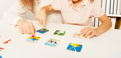 Na stole leży 6 obrazków. Przy stole siedzi dziecko i pokazuje na jeden z obrazków, obok niego osoba dorosła.