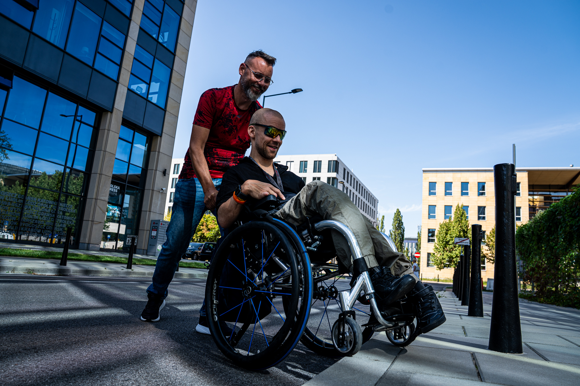 Mężczyzna na wózku inwalidzkim pokonuje przeszkodę na chodniku w przestrzeni miejskiej, a drugi mężczyzna pomaga mu, trzymając wózek od tyłu. W tle nowoczesne budynki biurowe i ulica w słoneczny dzień.