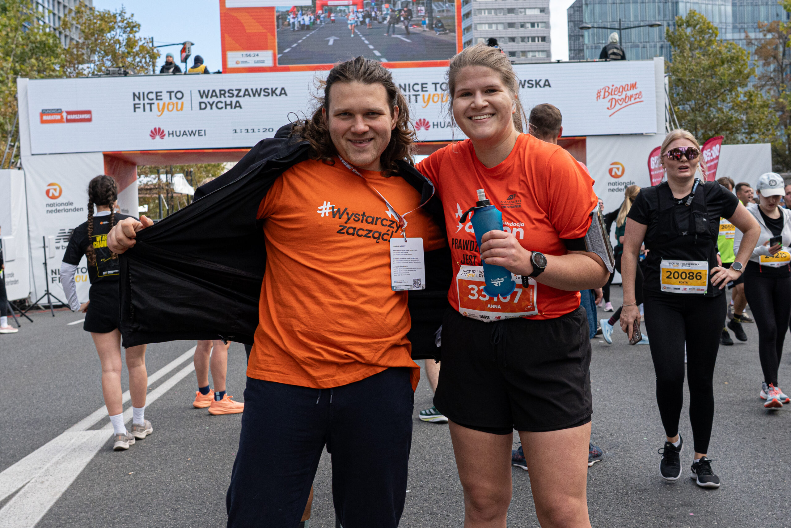 kobieta i mężczyzna pozują do zdjęcia ubrani w pomarańczowe koszulki Fundacji Avalon; w tle trasa maratonu warszawskiego