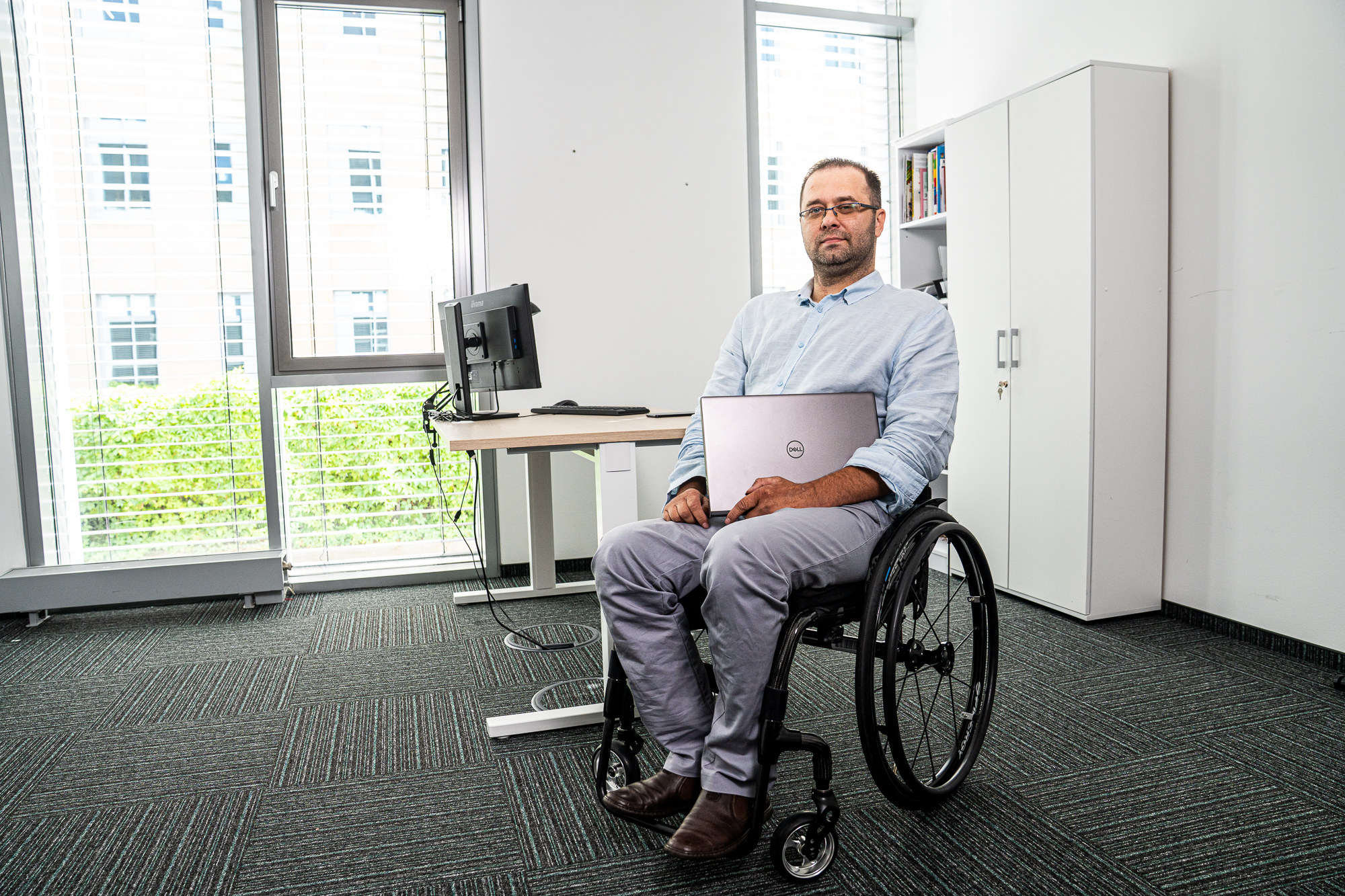 Mężczyzna na wózku z laptopem w tle biurko i szafa.