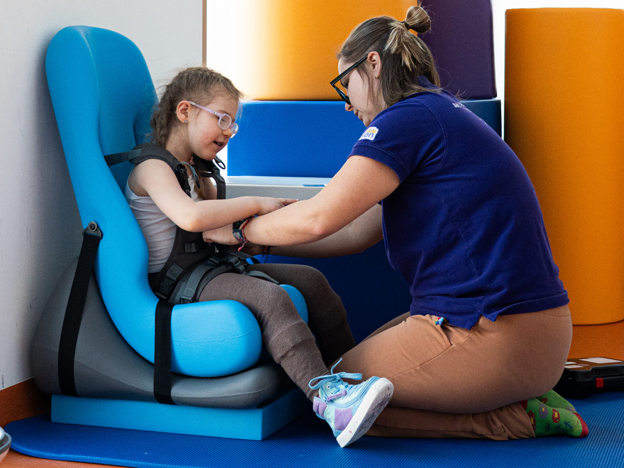 Dziewczynka siedzi w siedzisku. Obok fizjoterapeuta.