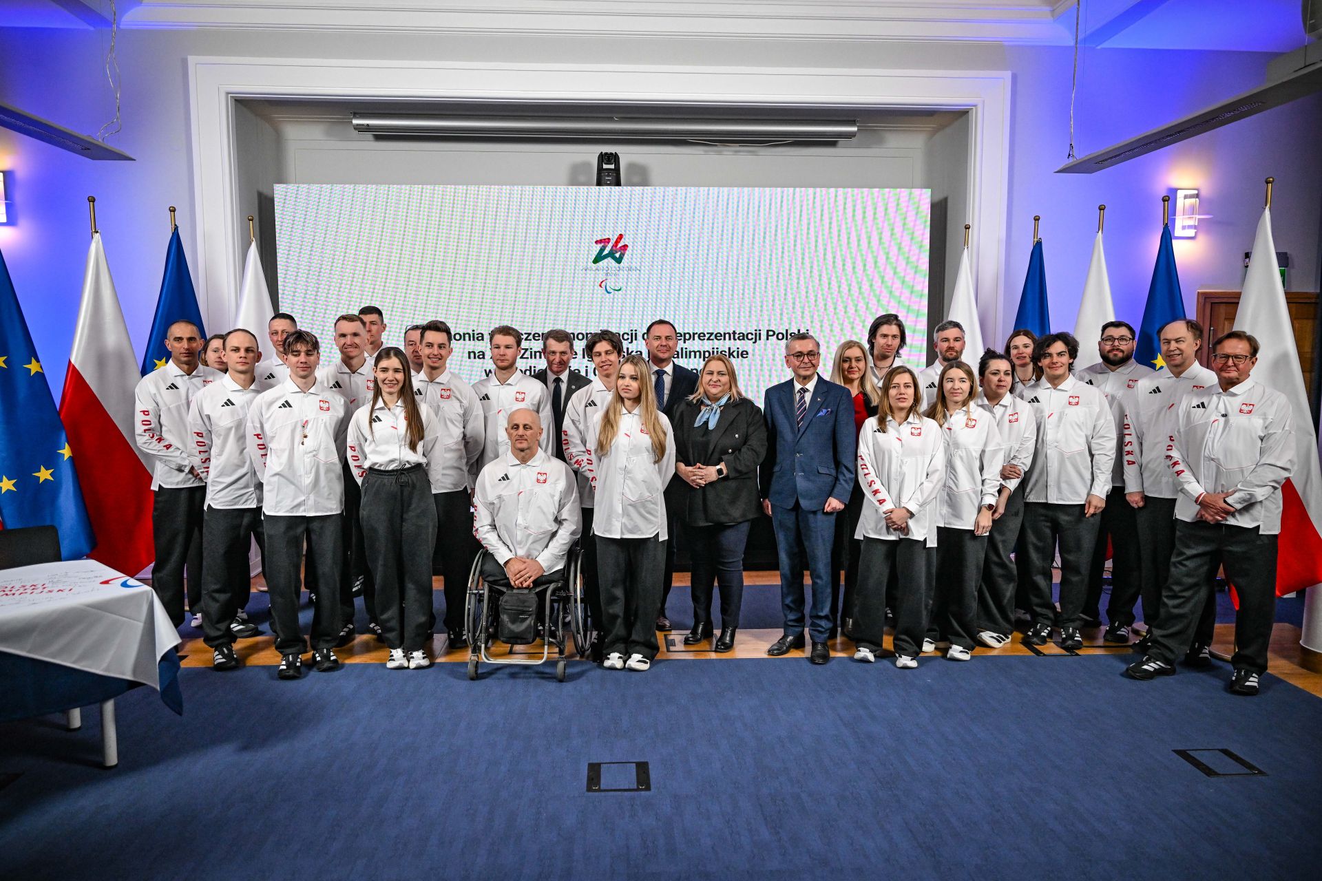 2026.02.18 Igrzyska Paralimpijskie
fot. Bartłomiej Zborowski wspólne zdjęcie grupowe sportowców nominowanych do Paralimpiady razem z gospodarzami wydarzenia i gośćmi; przestrzeń sali konferencyjnej, w tle projektor i flagi polski oraz Unii Europejskiej.