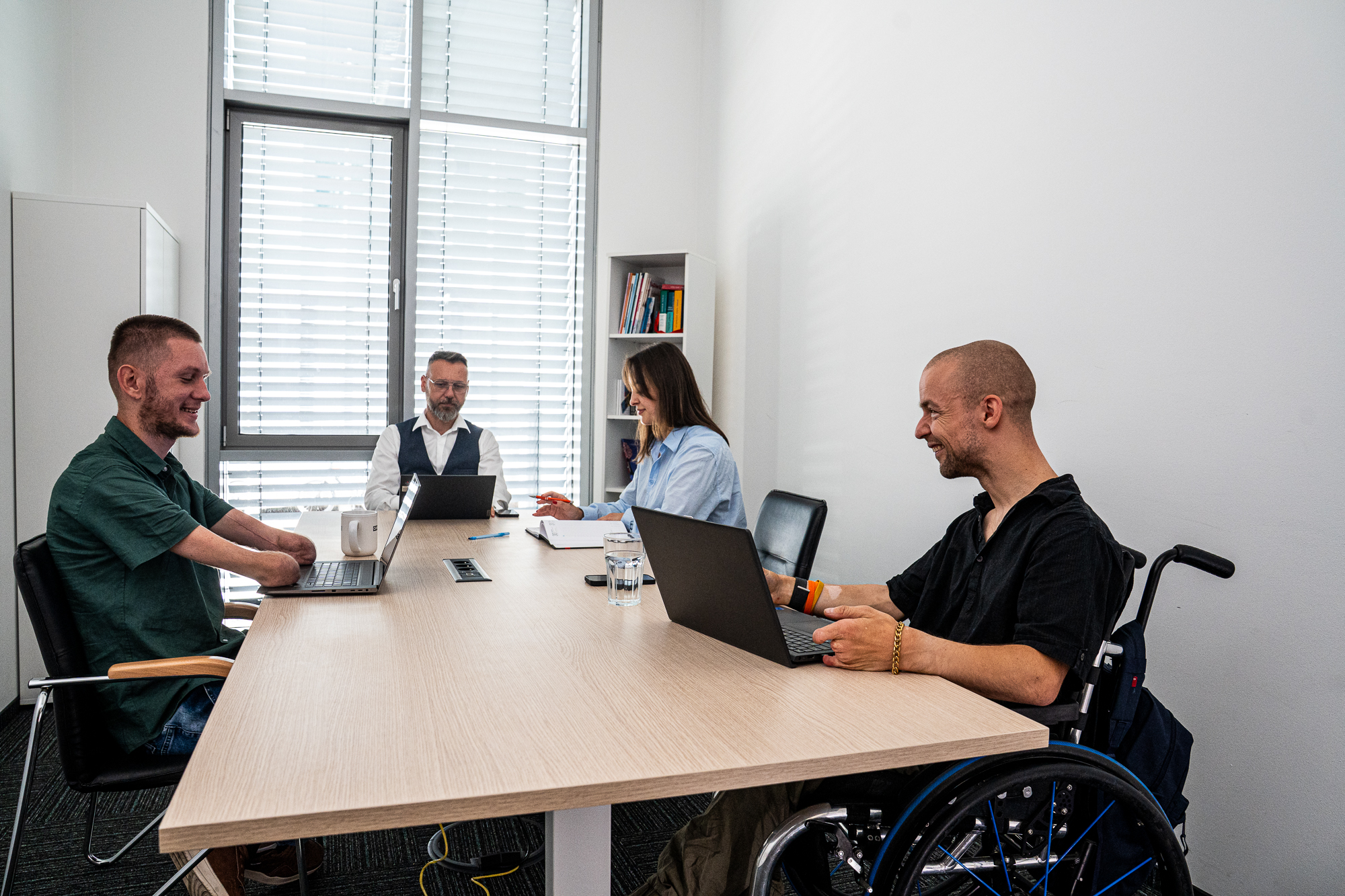 Trzech mężczyzn i kobieta siedzą wokół stołu w salce konferencyjnej. Przed nimi stoja laptopy. Jeden z męższyzn jest na wózku, a drugi ma ręce do łokcia.